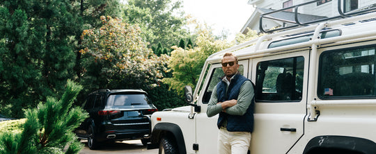 man in a sweater and vest leaning against a vehicle