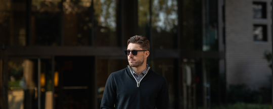 Man in sunglasses and a sweater, standing confidently outside a modern glass building."