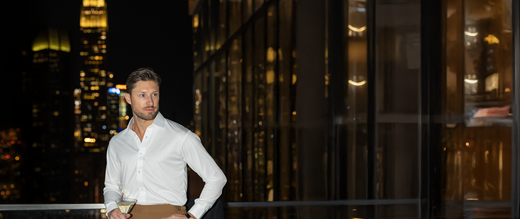 man in a city standing on a balcony in a white button down while holding a cocktail